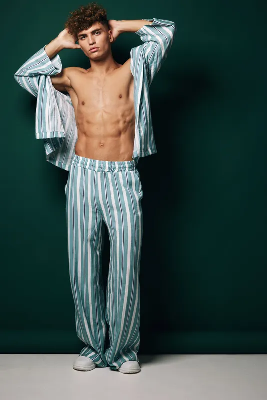Young man with curly hair posing shirtless in striped loose pants and open shirt against dark background, showing toned abs and confident expression in studio setting.