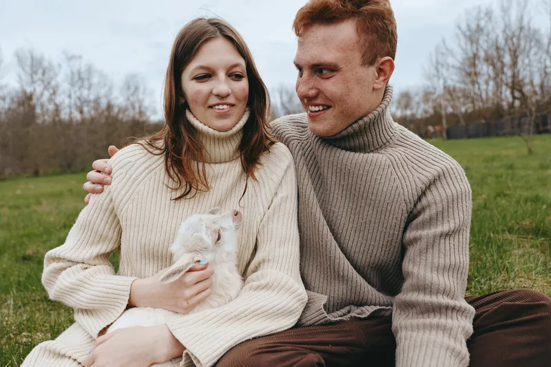 Cheerful couple outdoors, warm sweaters, soft sunlight, dog in their arms, relaxed laughter, casual moment, green field backdrop, natural colors, cozy atmosphere, nature