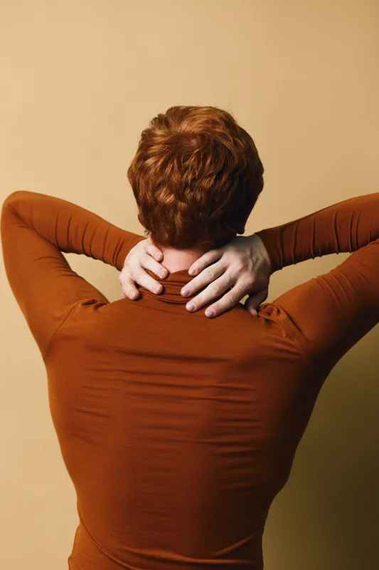 Person with red hair wearing brown long sleeve shirt holding neck and stretching against beige background. Back view with hands on neck and casual clothing.