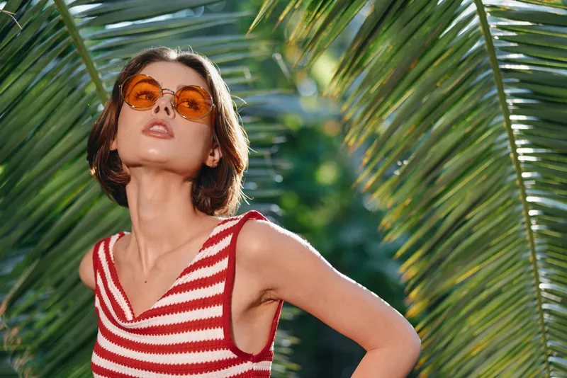 A stylish woman wearing a red and white striped top and orange sunglasses stands outdoors among tropical palm leaves, striking a confident pose under bright sunny skies, exuding bold summer vibe.