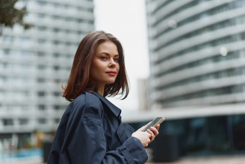 Young woman in a navy jacket stands in an urban setting, looking at her phone with a calm expression and a modern city backdrop.