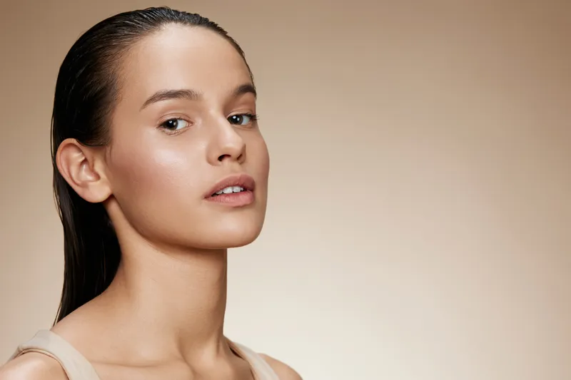 Beautiful young woman with natural makeup and wet hair, displaying a calm expression against a soft beige background, embodying beauty and confidence