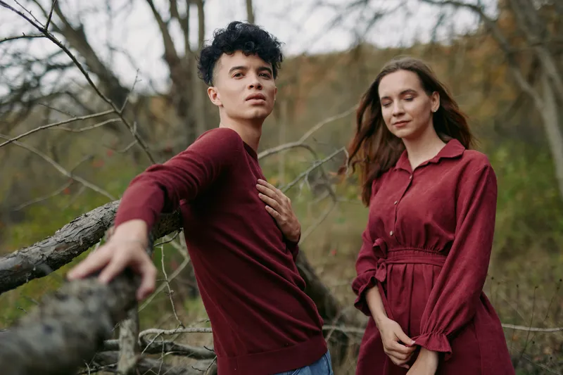 A candid portrait of a young couple in coordinated burgundy outfits, conveying authentic connection and genuine emotion as they stand against a rustic natural backdrop with soft light.