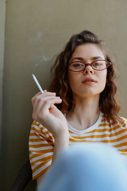 Casual woman at home sits against a neutral wall, smoking a cigarette, wearing a striped shirt and glasses, relaxed mood, thoughtful expression, intimate everyday moment for a cozy vibe.