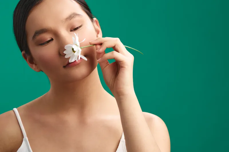 asian woman holds a white flower near her face, set against a bold green background. a serene, minimalist portrait that emphasizes natural beauty, calm mood, and modern simplicity.