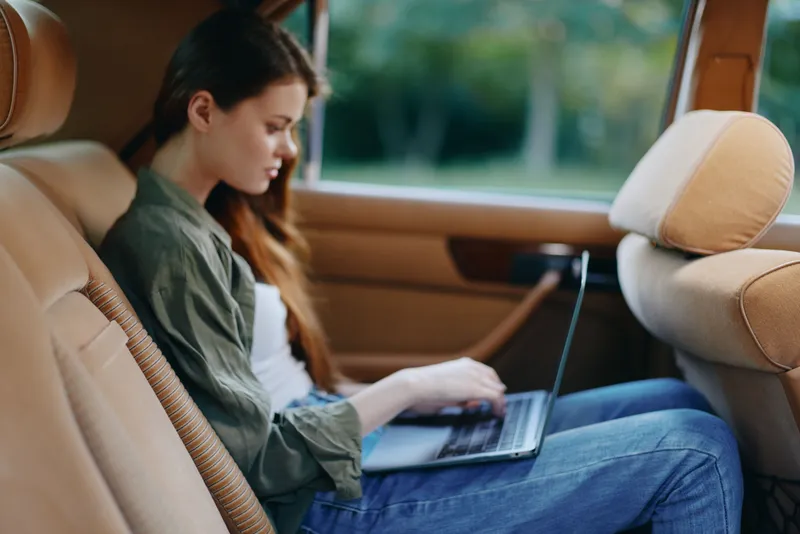 Young woman working on laptop in a stylish car interior, showcasing a blend of productivity and comfort with a focused expression.