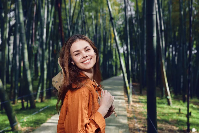 Woman smile backpack path forest trees nature outdoors portrait of a happy traveler walking a bamboo trail in green sunlight, casual orange jacket, young beauty enjoying a peaceful journey.
