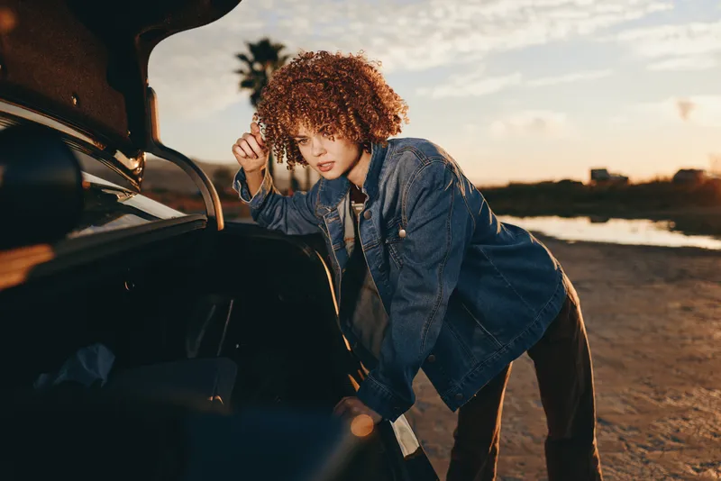 Woman with curly hair wearing denim, leaning by car door on an outdoor road at sunset, lifestyle travel mood, wind in hair, warm golden light, candid, evening sky glow, warm wind