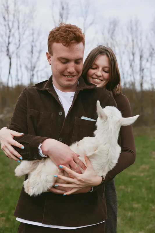 Smiling couple outdoors cuddling a white goat kid in a grassy field, warm interaction, soft natural light, rustic countryside mood, joyful companionship.