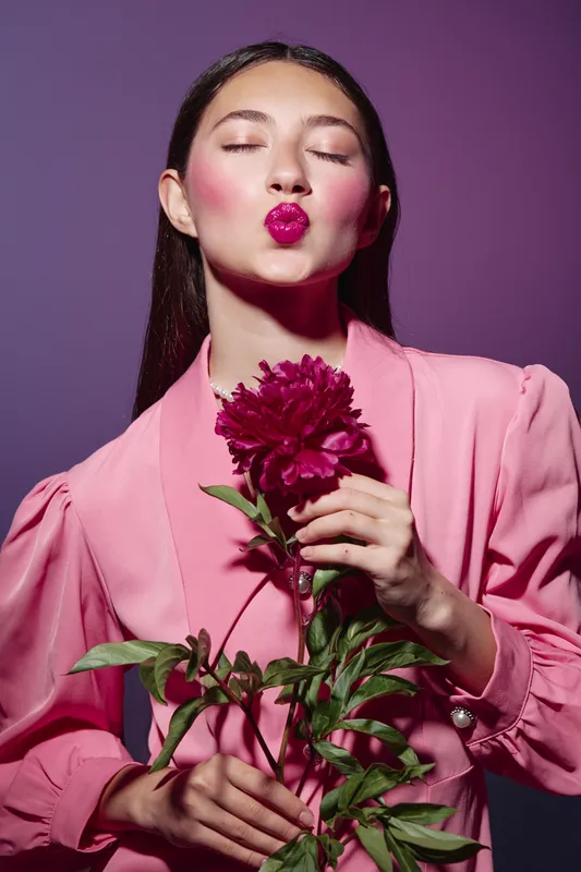 Young woman wearing pastel pink jacket holds red flower and blows kiss with bright makeup on purple background. Concept for beauty creative fashion style and feminine elegance.