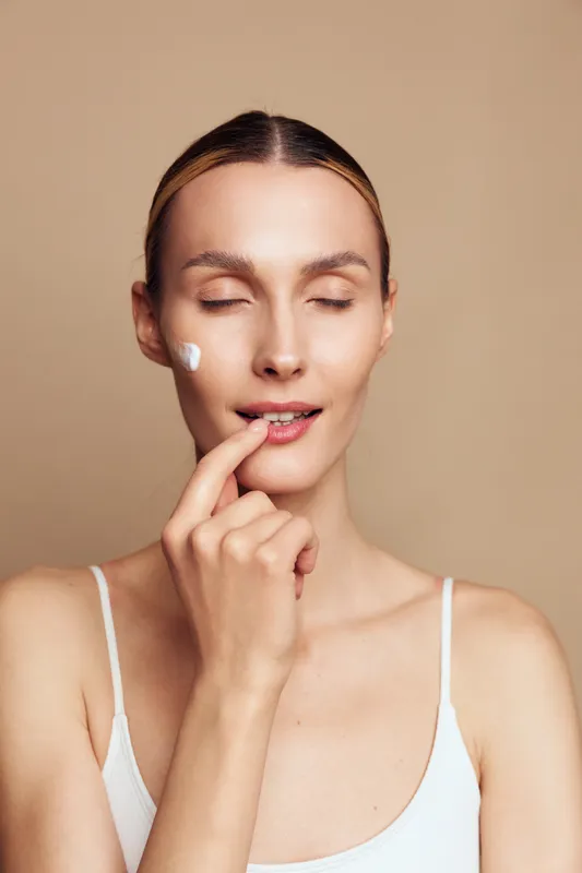 Young woman with closed eyes applies facial cream on her cheek with a gentle touch against a neutral beige background in a skincare routine moment.