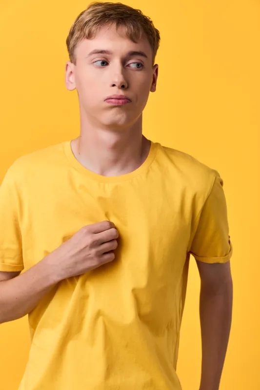 Man in a yellow shirt stands before a bright yellow backdrop, adjusting his collar with a thoughtful gaze, studio lighting emphasizing casual fashion and mood for lifestyle or catalog use