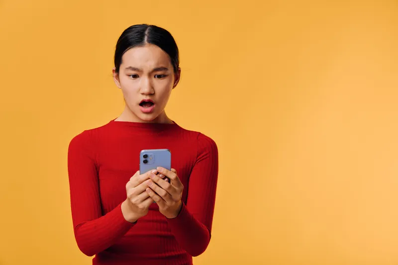 Young woman in red sweater looking surprised with smartphone in hands on yellow background for communication and digital reaction. Emotion, technology and mobile device concept.