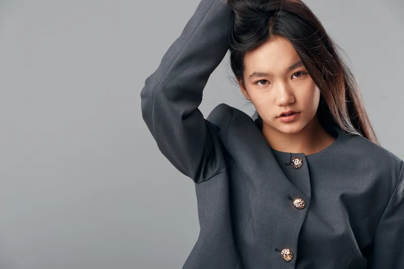 Young woman with long hair wearing dark gray buttoned jacket posing with hand touching hair against neutral gray background for fashion or portrait concept.