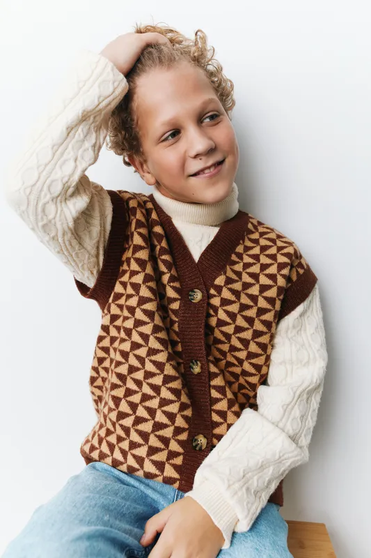 Portrait of a cheerful child wearing a geometric patterned brown vest over a cream knit sweater, posing relaxed against a clean white background with a gentle smile.