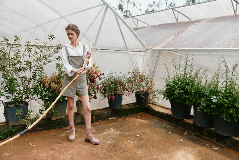 Young woman in a greenhouse gardening with a rake, wearing casual clothing, promoting healthy living and sustainability during springtime