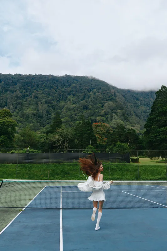 Young female model in a white dress playfully twirling on a tennis court surrounded by lush green mountains, conveying joy and freedom