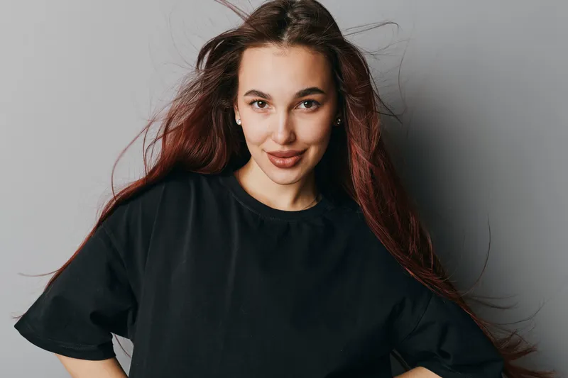 Young woman with long hair and natural makeup wearing black t shirt posing against gray background looking confident and smiling with hands on hips in casual style portrait indoors.