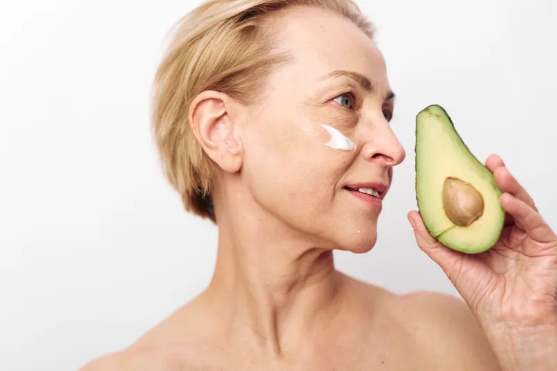 Mature woman with short blonde hair applies white cream on her cheek holding fresh avocado half near face. Skincare concept with natural beauty, healthy lifestyle, and organic cosmetics.