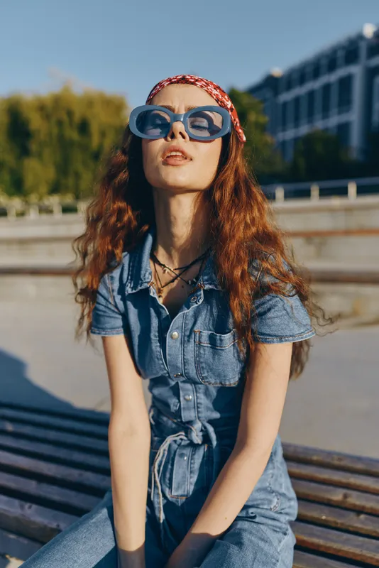stylish woman wearing denim jumpsuit and oversized sunglasses posing outdoors with headscarf and curly red hair in bright sunny urban setting