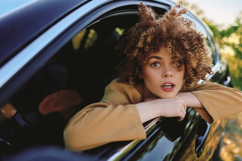 Cheerful woman leaning out of a car window, sunlit breeze, casual outfit, candid lifestyle moment, retro film tones, natural colors, relaxed playful expression in warm evening light and soft focus,