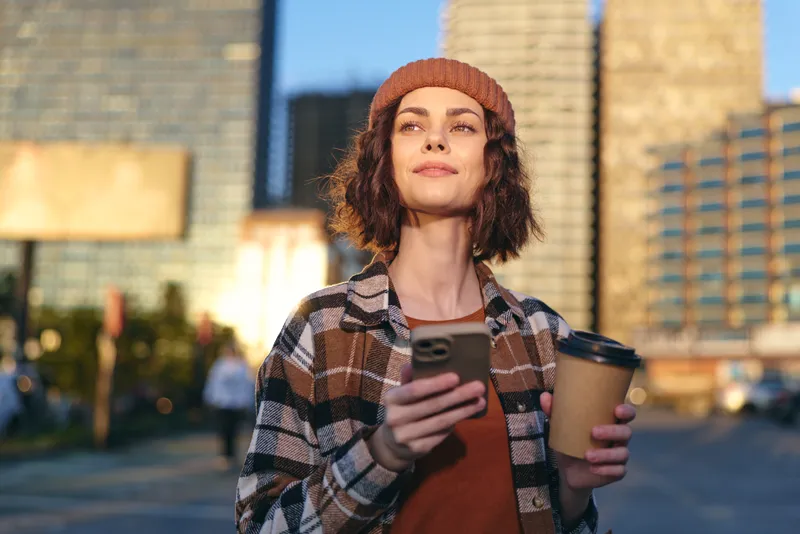 Woman with coffee and smartphone in city street, wearing beanie and plaid jacket, sunlight on her face. Candid authenticity, golden hour glow, mindful living and emotional storytelling urban