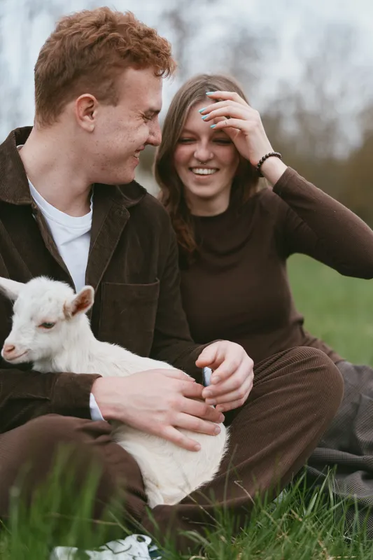 Couple and goat kid in a rural field enjoying a warm outdoors lifestyle moment, smiling, gentle touch, casual clothing, grass underfoot, natural light, candid affection, nature