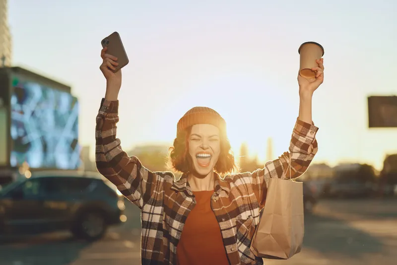 Woman joyful with coffee and phone, shopping bag raised in city sunlight, smile and excitement in golden hour glow; candid lifestyle portrait highlighting authenticity, mindful living and emotional