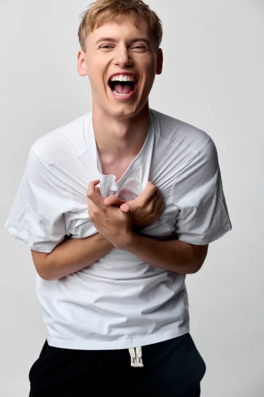 Laughing male in white shirt, casual style, grabbing chest with a wide grin, energetic and candid mood capturing joy and carefree emotion