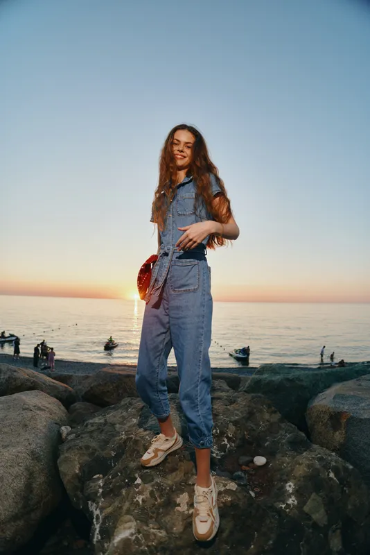 Lifestyle woman wearing modern boho-western denim street style outfit stands on rocks by the sea during golden hour with film color, relaxed and confident appearance.