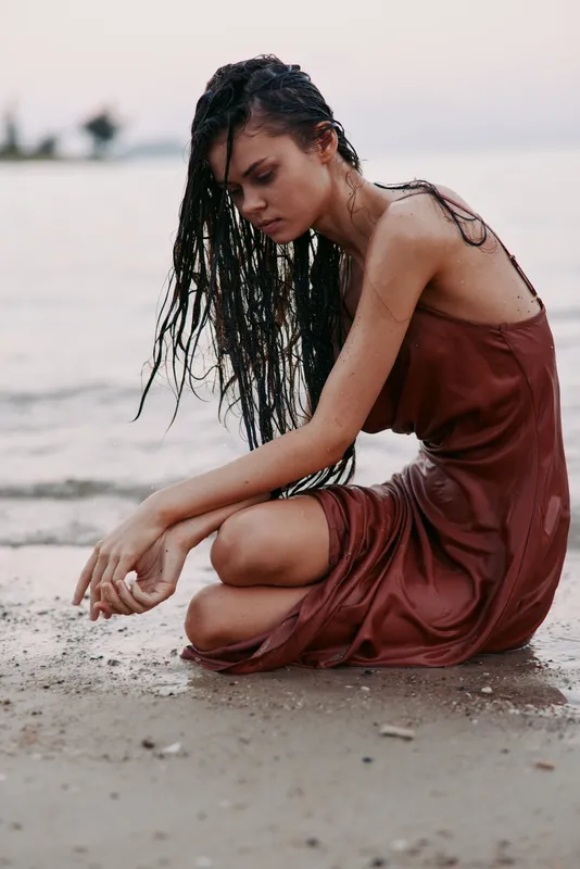 A contemplative woman kneels on a beach wearing a rust dress, wet hair cascading, soft light, calm mood, and subtle reflection creating an intimate coastal portrait.