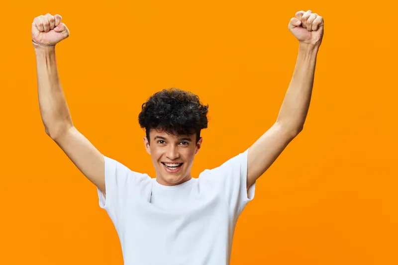 young man celebrating with raised arms, smiling enthusiastically against bright orange background, casual white shirt, joyful and energetic expression