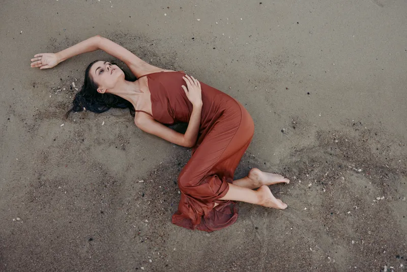 Relaxed woman lying on sandy beach wearing rust dress, enjoying calm moment with closed eyes and peaceful expression in natural outdoor setting.
