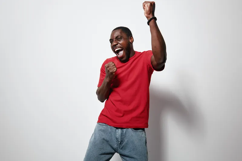 African American man with dark skin wearing red t shirt and jeans celebrating with raised fist and joyful expression on white background. Casual clothing and happiness.