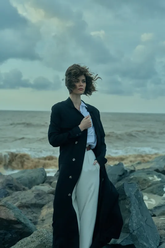 Fashion silhouette along a windswept shoreline, long black coat over white garments, dramatic clouds over the ocean, edgy styling for editorial or campaign visuals.