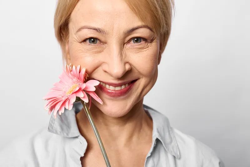 Woman smiling portrait flower mature closeup studio pink blossom on stick near mouth, soft light, relaxed eyes, gray shirt, white background