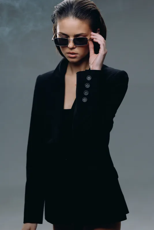 Stylish woman in black blazer wearing sunglasses with confident pose and moody gray background