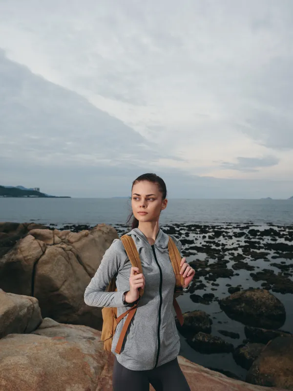 Young woman exploring rocky coastline with a backpack, wearing a gray sporty jacket, facing the sea, showcasing adventure and outdoor lifestyle