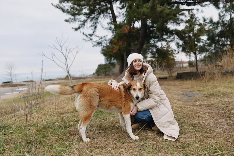 Woman in a warm coat hugging a friendly dog in an outdoor park with grass and a calm lake in the background for a gentle companionship moment