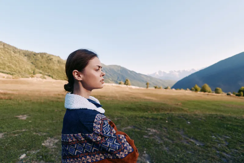 Woman portrait in mountains and meadow, outdoors nature scene with thoughtful expression, standing in a landscape field wearing a patterned sweater, serene travel solitude mood.