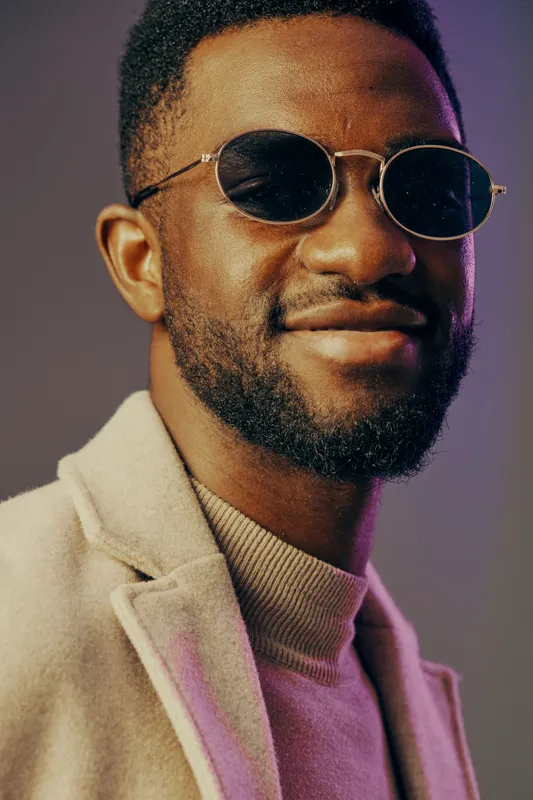 Stylish African man in sunglasses, showing confidence with a warm smile, dressed in a cozy sweater and coat, against a colorful backdrop