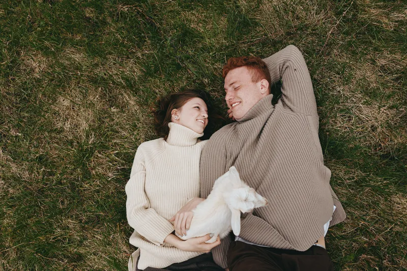 An affectionate couple lying on grass with a curious goat kid between them, enjoying a sunny outdoor moment. A candid lifestyle scene highlighting companionship, nature, and animal bonding.