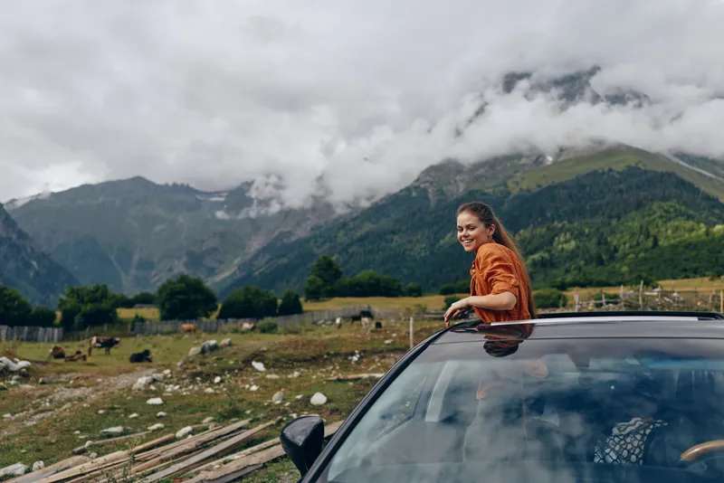 Female traveler leans from a car window amid a scenic mountain landscape, smiling at the camera as clouds drift over green valleys, capturing a moment of outdoor adventure.