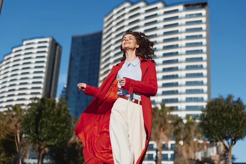 Young woman in a redcoat and whitepants walks outdoors in an urban plaza, smiling toward the sun as tall buildings rise behind her, a carefree, breezy moment.
