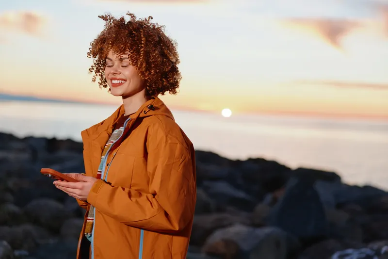 Smiling woman with curly hair wearing an orange jacket by the beach at sunset, outdoors lifestyle scene, golden sky and sea waves in soft focus, calm travel mood, natural light