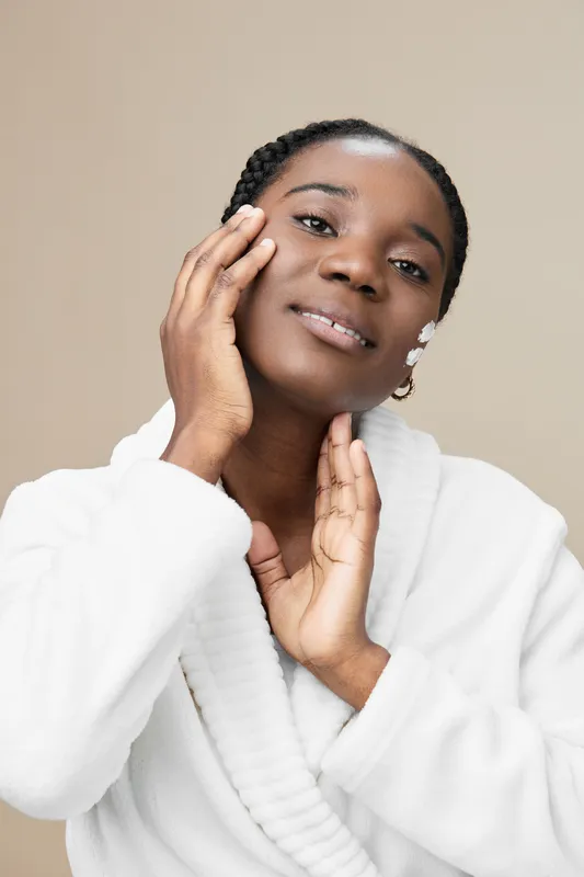 Young Black woman in a white robe smiling softly, showcasing skincare routine on a neutral background with a warm color palette