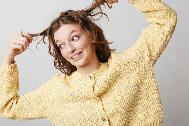 Young woman in yellow knitted sweater plays with her hair and smiles looking sideways on grey background. Concept of casual style, happiness, and carefree mood.