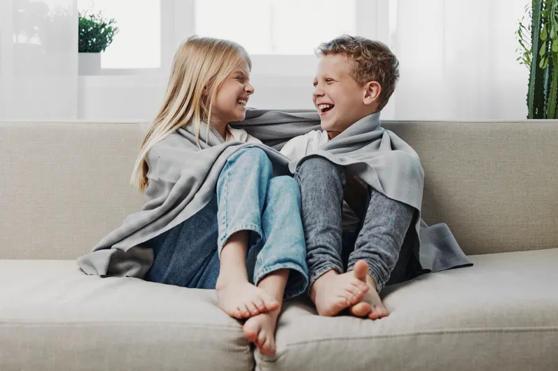 Two happy children wrapped in grey blankets sit barefoot on a light sofa, laughing and looking at each other in a bright cozy living room. Concept of warmth, friendship, and childhood joy.