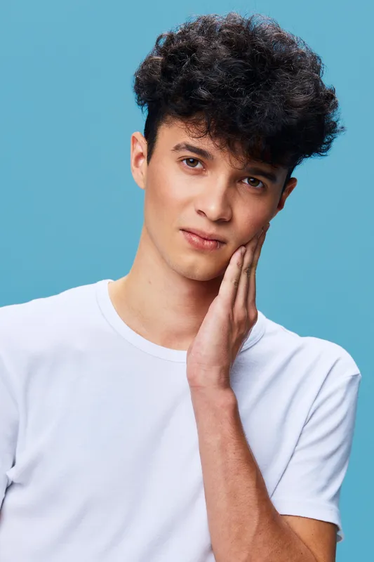 Young man with curly hair wearing white t-shirt touching his face against blue background, expressing calm and thoughtful mood with casual style.