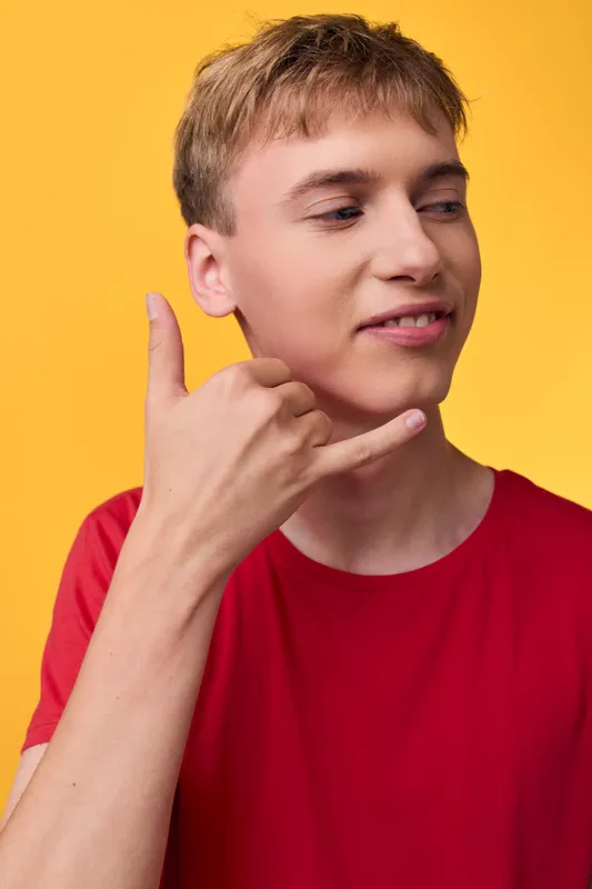 Young man in a red shirt makes a call me hand gesture against a bright yellow background, showing a casual pose and friendly communication for social media or advertising concepts